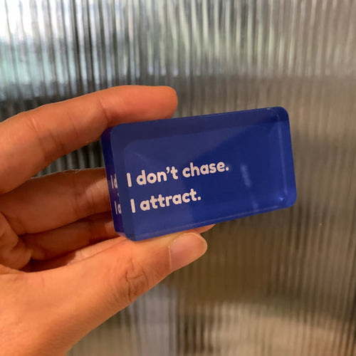 Hand holding a blue fridge magnet with text " I don't chase, I attract."