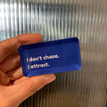 Hand holding a blue fridge magnet with text " I don't chase, I attract."