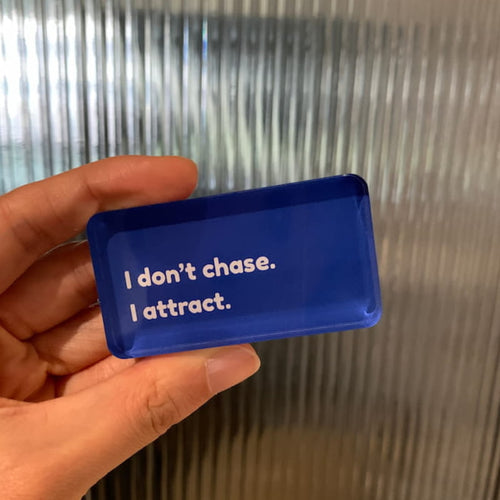 Hand holding a blue fridge magnet with text " I don't chase, I attract."
